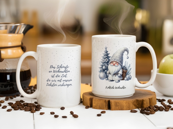 Zwei weiße Weihnachtstassen stehen auf einem weißen Tisch, umgeben von Kaffeebohnen und einer Kaffeemaschine im Hintergrund. Die linke Tasse zeigt den Text "Das Schönste an Weihnachten ist die Zeit, die ihr mit euren Liebsten verbringt." Die rechte Tasse
